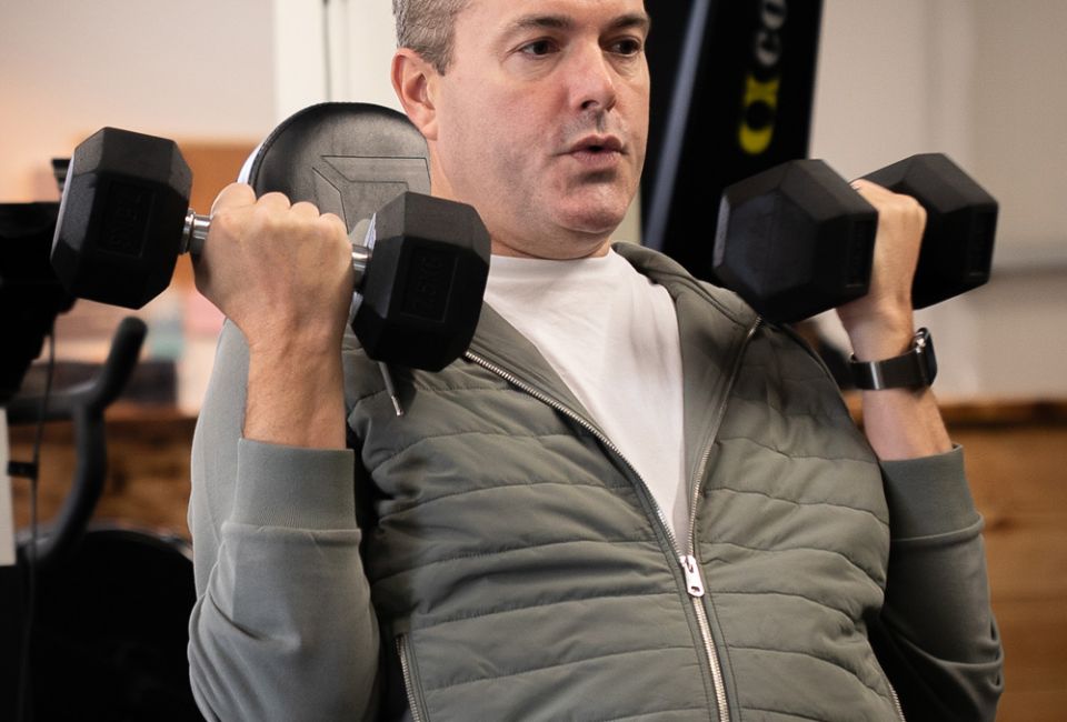 A man in a gym performs shoulder presses with dumbbells. He is wearing a gray tracksuit and appears focused and determined, conveying a sense of concentration.