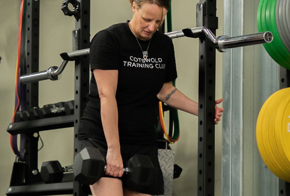 A woman in a "Cotswold Training Club" shirt holds a dumbbell near weight racks in a gym. The setting is focused and energetic.