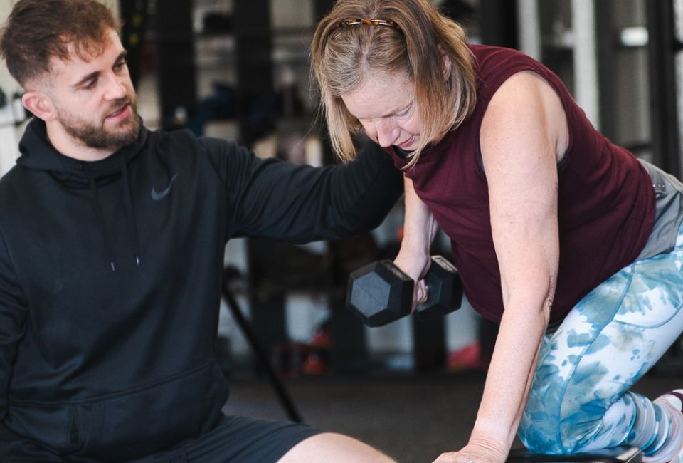 A woman in workout clothes performs a dumbbell row on a gym bench, assisted by a male trainer in black sportswear, conveying encouragement and focus.
